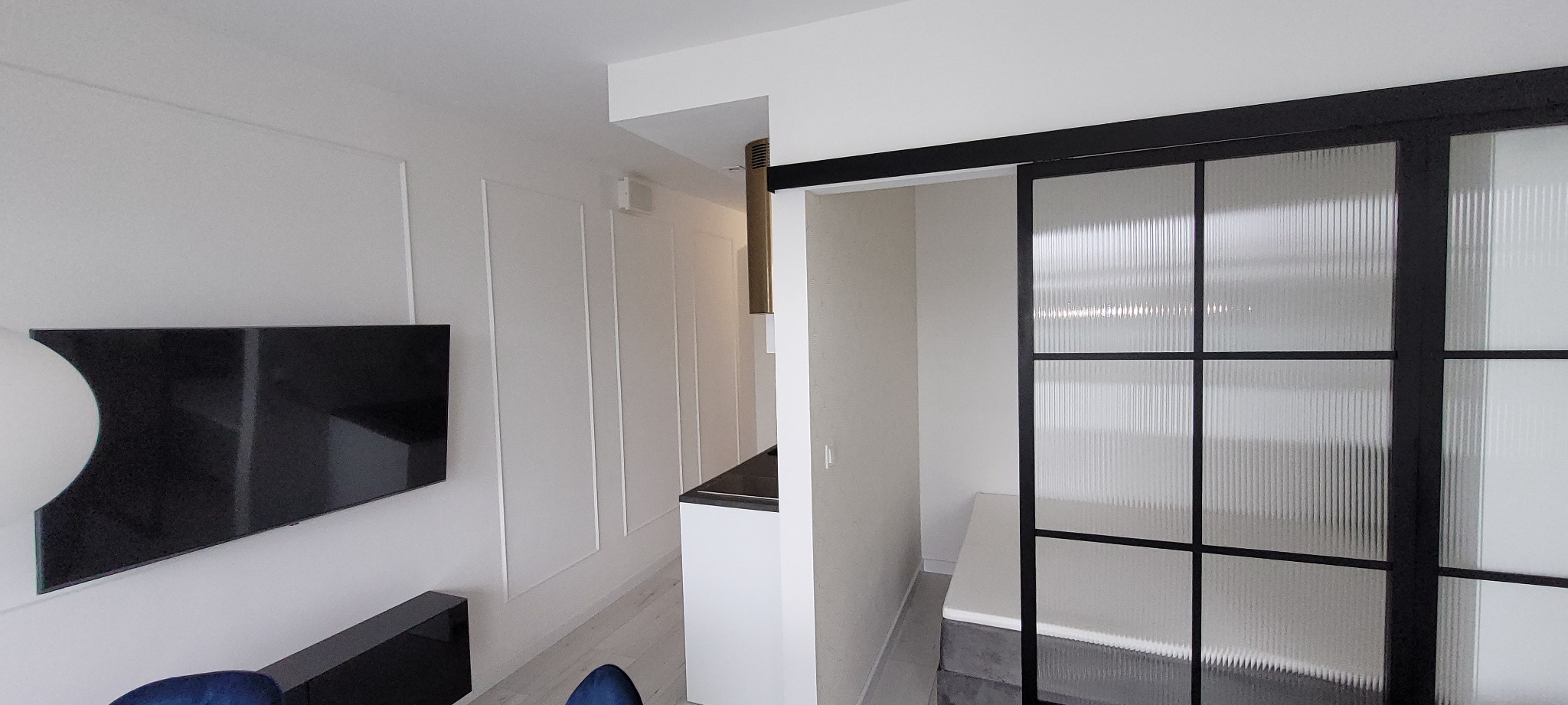 Nowoczesne wnętrze z czarnym telewizorem na ścianie z dekoracyjnymi listwami, widok na kuchnię z złotym okapem i przesuwne drzwi z ryflowanego szkła w czarnej ramie.