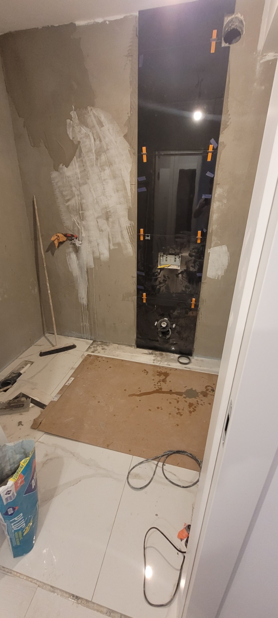 Łazienka w trakcie remontu: ściany częściowo pokryte gładzią, zamontowany stelaż podtynkowy WC z czarną płytą, podłoga wyłożona białymi płytkami, widoczne narzędzia i materiały budowlane.