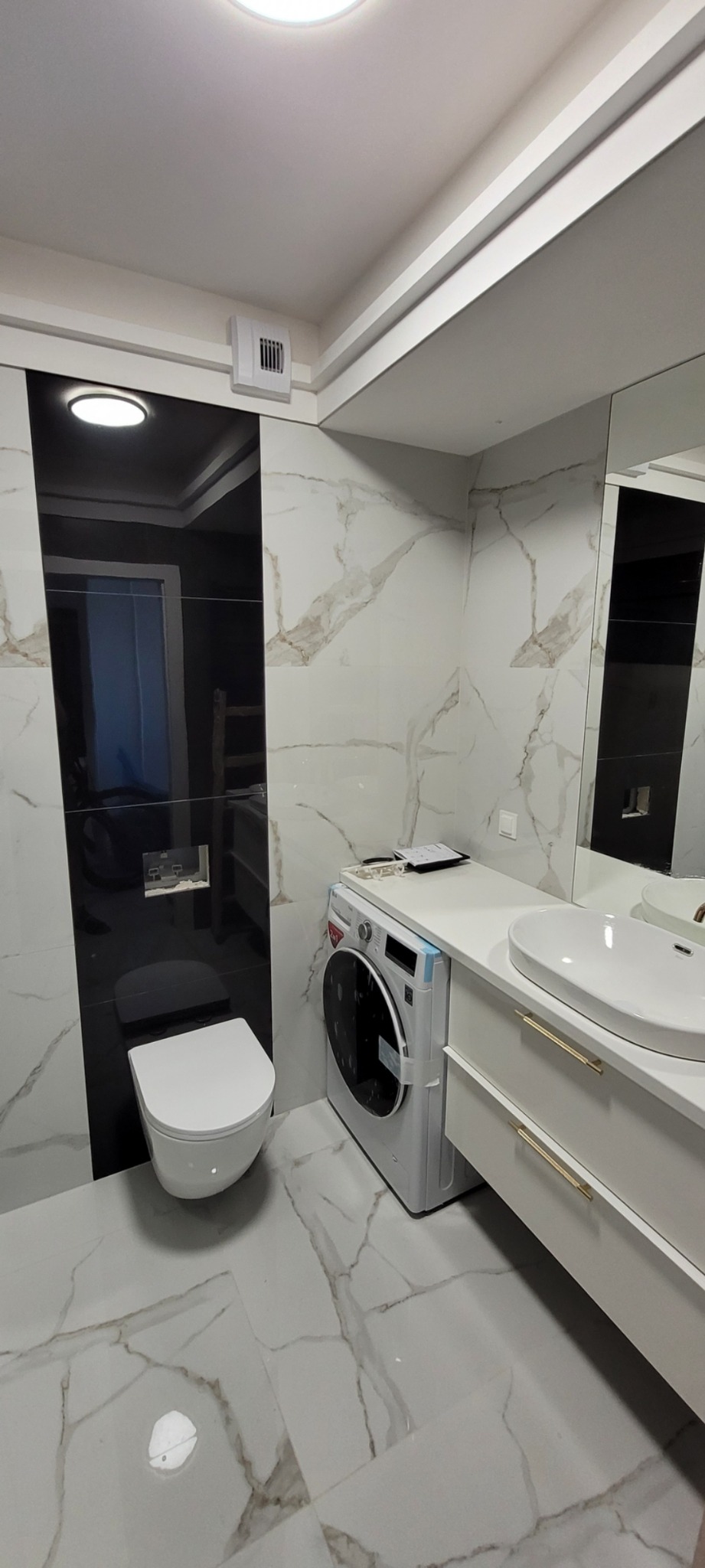 Nowoczesna łazienka z biało-czarnymi płytkami imitującymi marmur, wisząca toaleta, pralka pod blatem i umywalką z szafką z szufladami w kolorze białym.