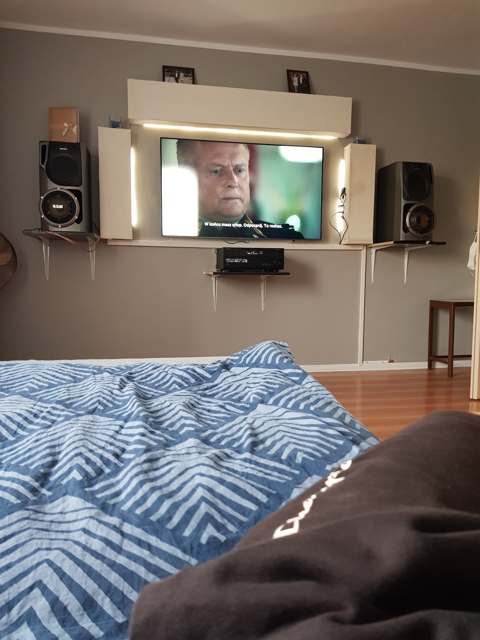 Telewizor z podświetleniem LED zamontowany na szarej ścianie, pod nim półka z dekoderem, po bokach głośniki na półkach z nietypowymi wspornikami, w tle fragment sypialni z łóżkiem w niebieskiej...