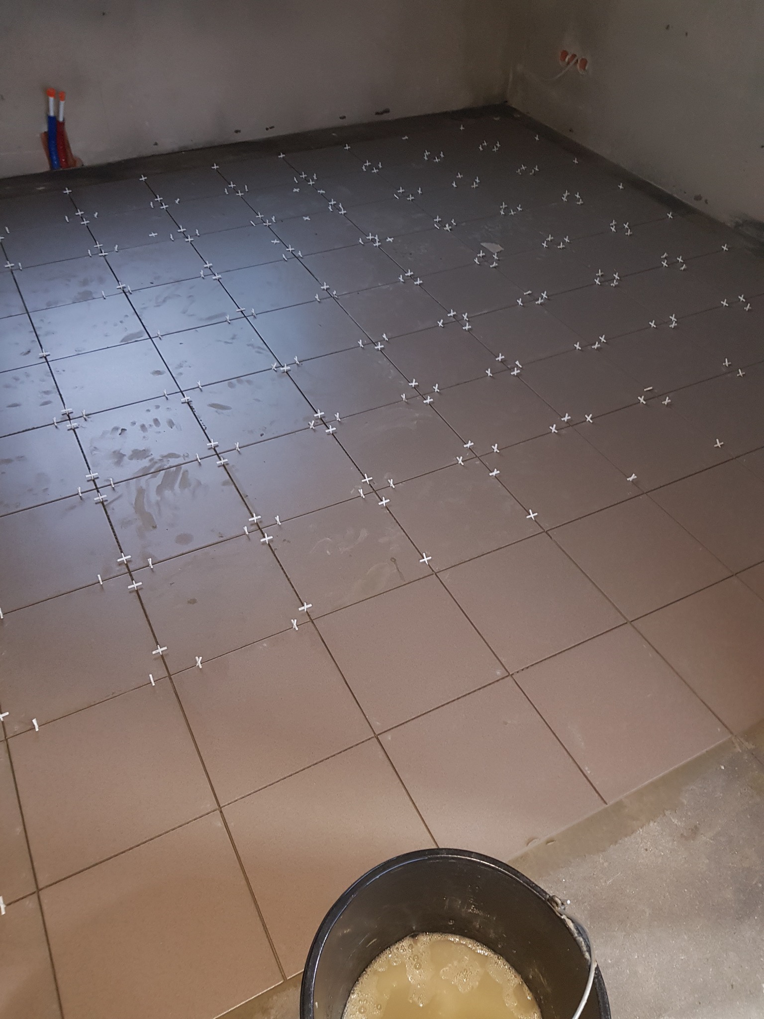 Układanie brązowych płytek podłogowych z krzyżykami dystansowymi w Kołobrzegu, widoczne rury instalacyjne na ścianie i wiadro z zaprawą.