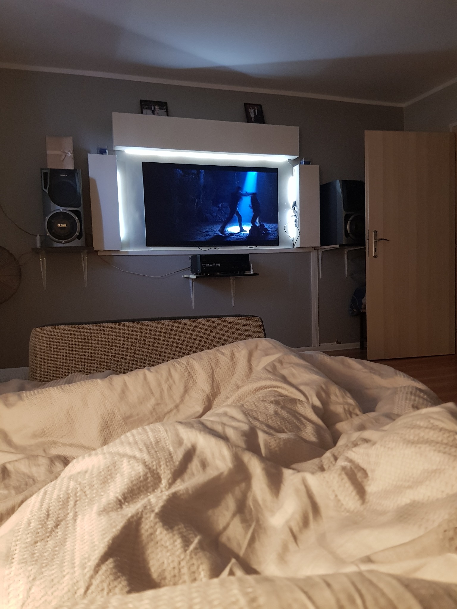 Telewizor na ścianie z podświetleniem LED, głośniki na półkach, widok z łóżka z pościelą.