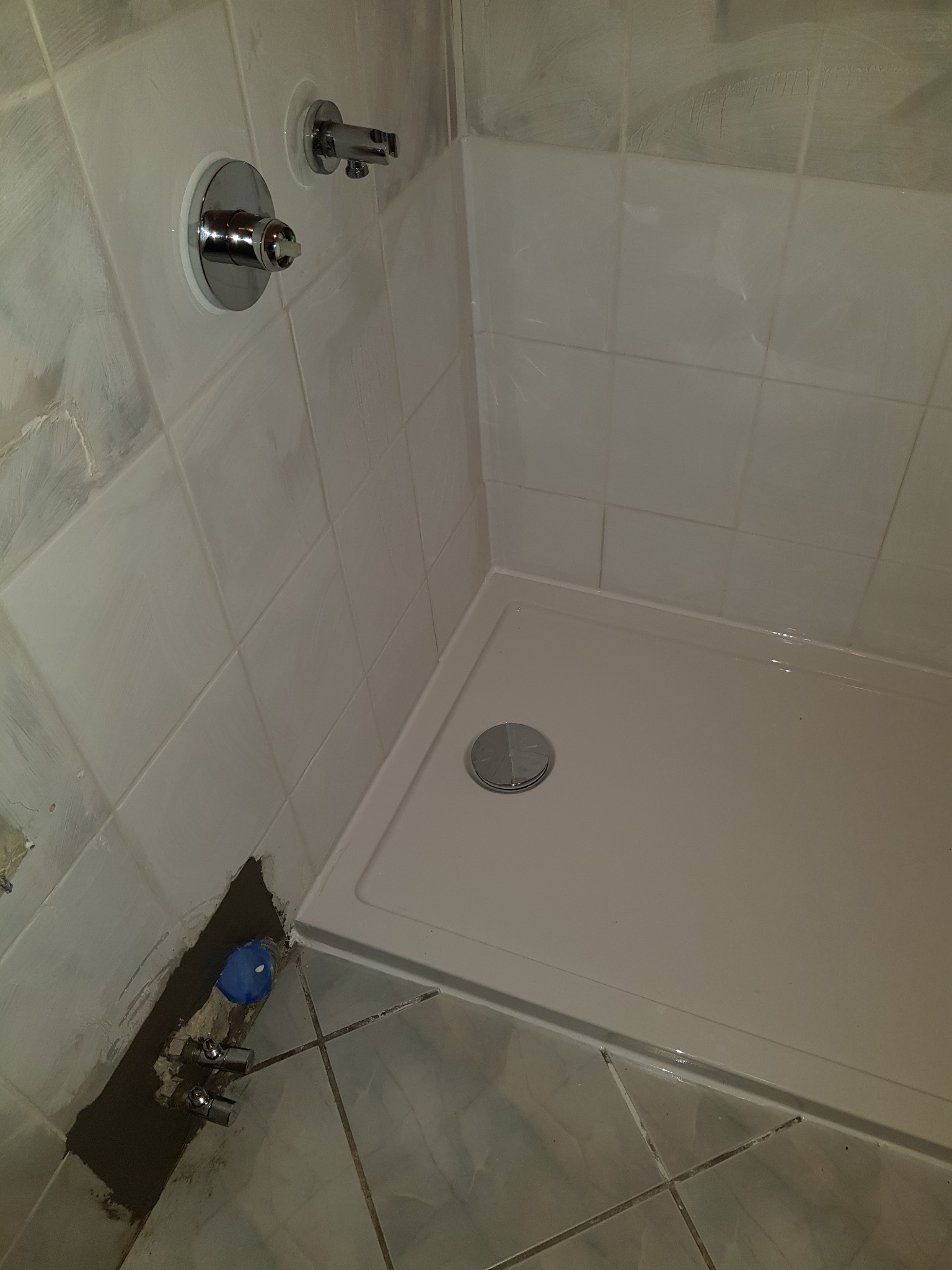 Narożnik kabiny prysznicowej z brodzikiem, widoczne chromowane zawory i bateria, surowe tynki przy podłodze, białe płytki częściowo pomalowane farbą.