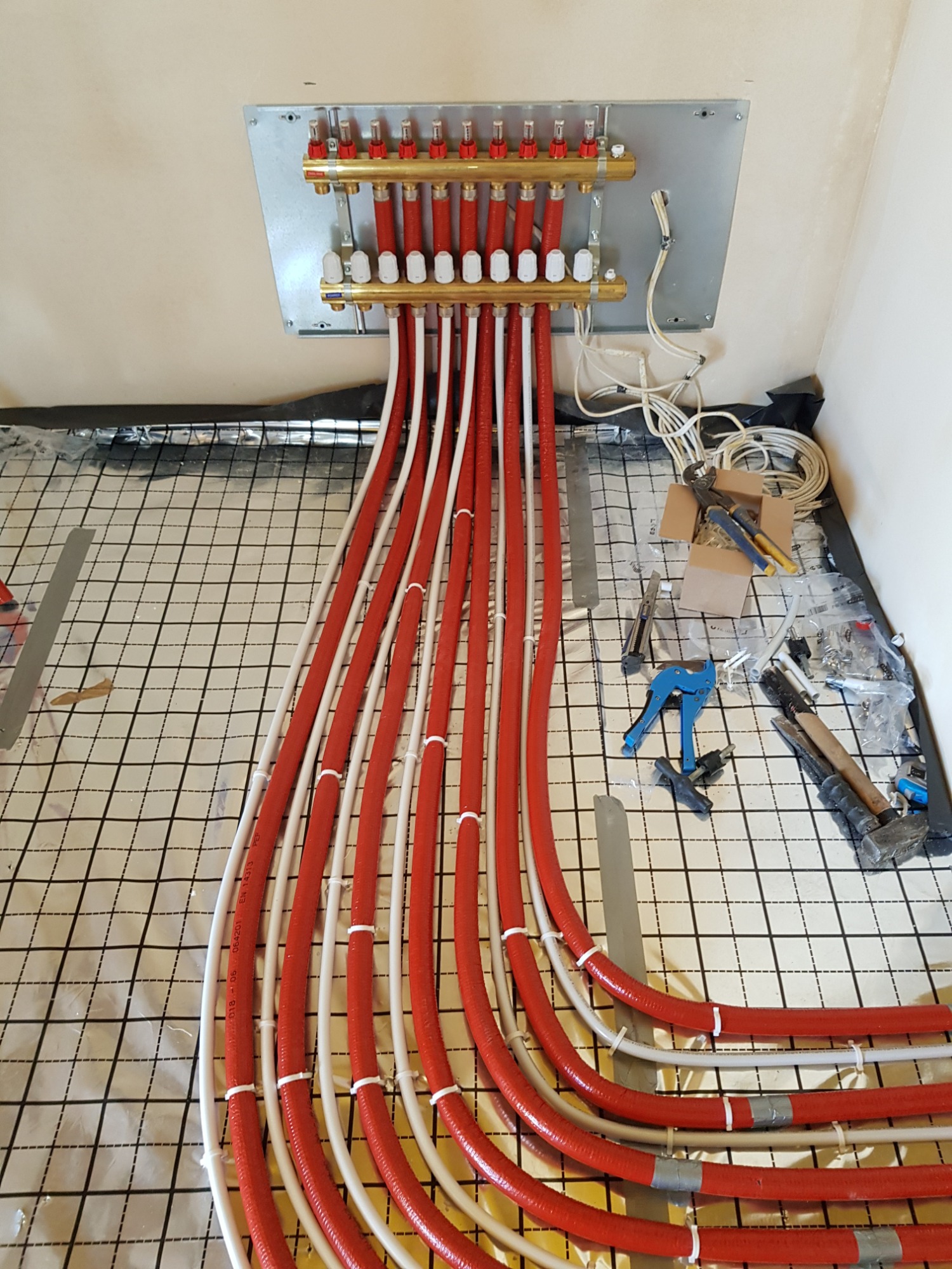 Rozdzielacz ogrzewania podłogowego z rurami w kolorze czerwonym i białym, przymocowanymi do folii z nadrukowaną siatką, widoczne narzędzia i akcesoria instalacyjne.