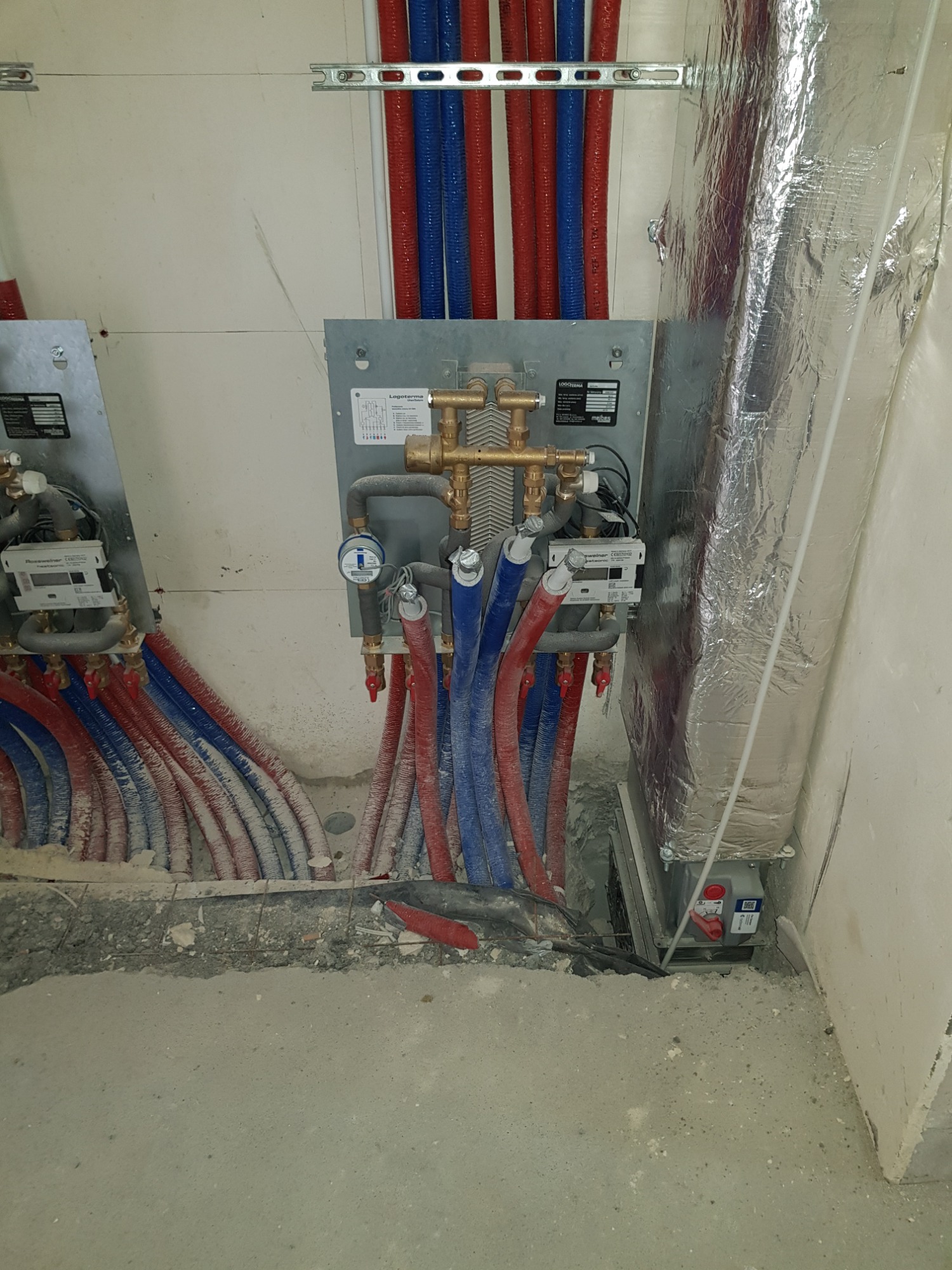 Rozdzielacz instalacji grzewczej z podłączonymi rurami w kolorze czerwonym i niebieskim, zamontowany na ścianie w surowym stanie, widoczne liczniki i izolacja termiczna kanału wentylacyjnego...
