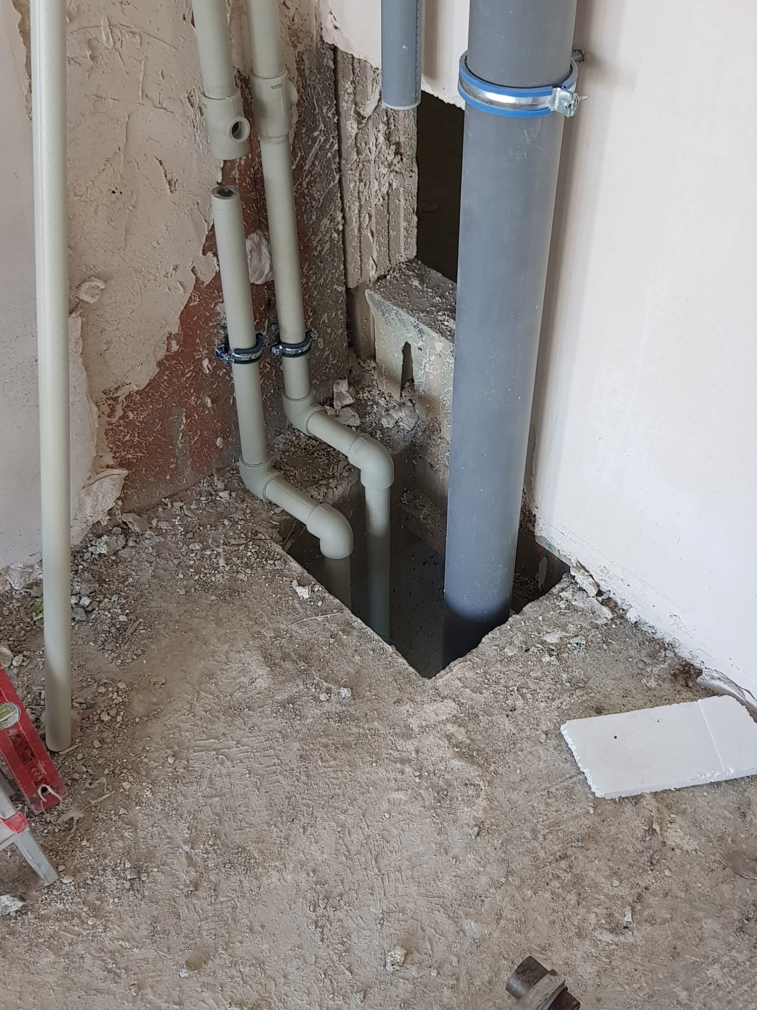 Widok na fragment ściany i podłogi w trakcie prac instalacyjnych. Widoczne rury z tworzywa sztucznego, w tym jedna szara pionowa rura zamocowana obejmą z niebieskim akcentem, oraz fragment...