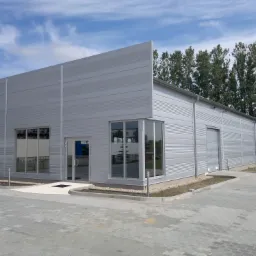 Gotowa hala przemysłowa z szarej blachy trapezowej, widoczne okna narożne i brama wjazdowa, kostka brukowa przed budynkiem, błękitne niebo z chmurami.