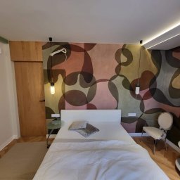 Lukasz Pierzchala - Sypialnia z łóżkiem na tle ściany z abstrakcyjnym wzorem. Wiszące lampy po bokach łóżka, drewniane drzwi i regał w tle. Jasne, nowoczesne wnętrze.