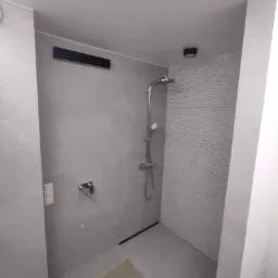 Nowoczesna łazienka w odcieniach szarości z prysznicem typu walk-in, wykończona drobną mozaiką na jednej ze ścian, widoczne elementy armatury i oświetlenie sufitowe.