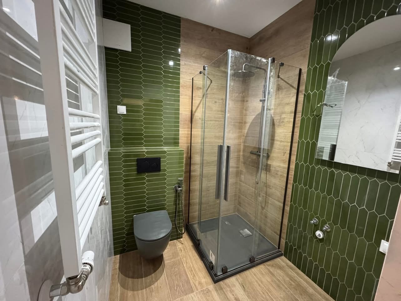 Nowoczesna łazienka z kabiną prysznicową, zielonymi płytkami w kształcie rombów i drewnopodobną podłogą. Grzejnik drabinkowy i szara toaleta podwieszana.
