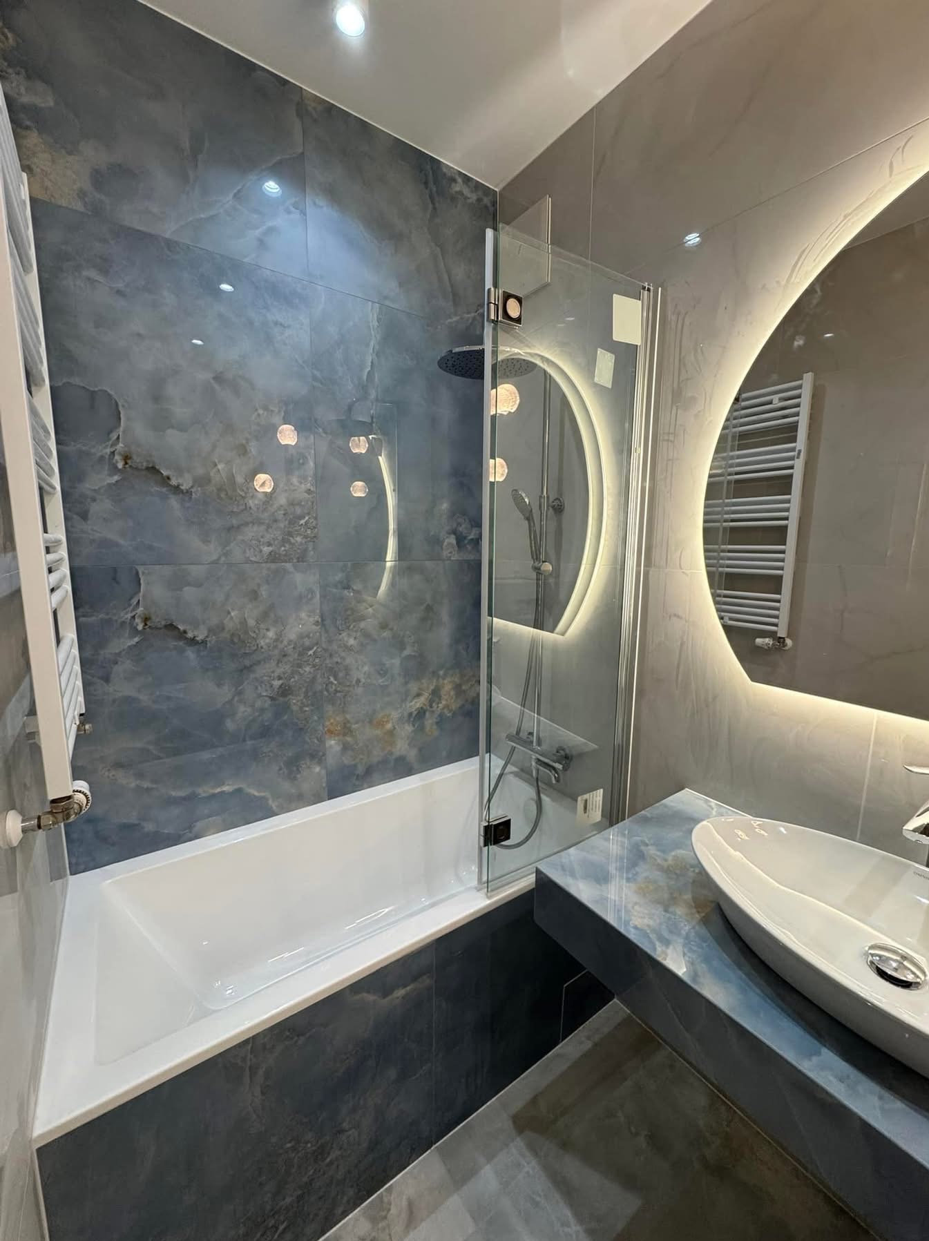 Wykończona łazienka z wanną obudowaną płytkami imitującymi marmur, szklanym parawanem, umywalką nablatową i okrągłym lustrem z podświetleniem LED.