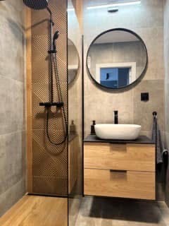 Nowoczesna łazienka z kabiną typu walk-in, okrągłym lustrem i drewnianą szafką pod umywalką. Uwagę zwraca panel dekoracyjny pod prysznicem.