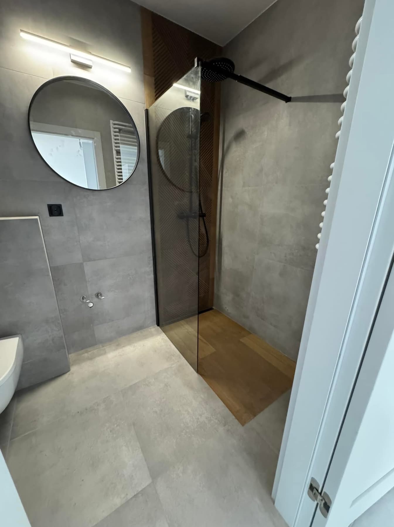 Nowoczesna łazienka z kabiną typu walk-in, drewnianym akcentem w strefie prysznica i okrągłym lustrem. Szare płytki na ścianach i podłodze. Minimalistyczny design.
