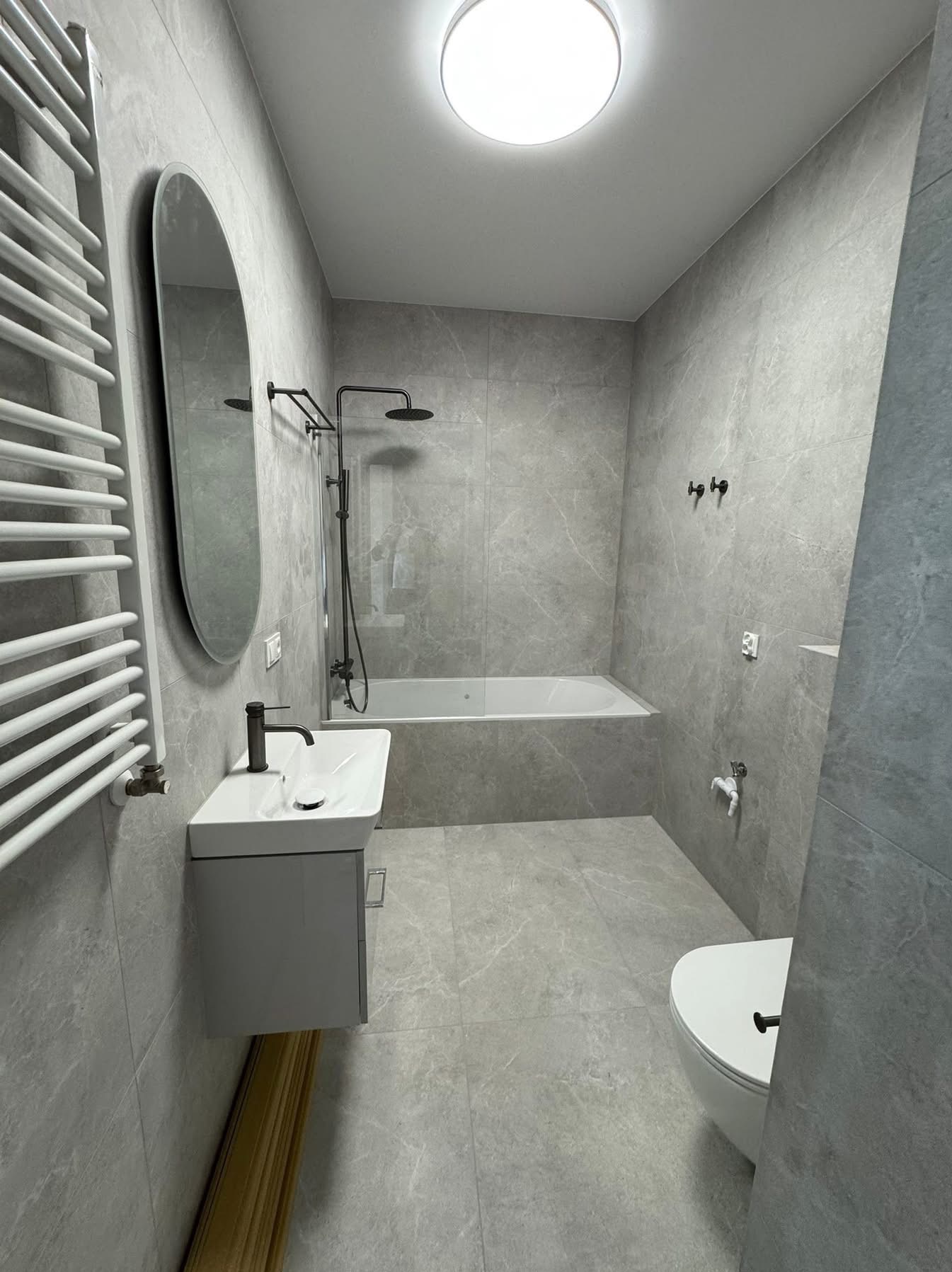 Nowoczesna łazienka w szarościach: owalne lustro, biała umywalka z szafką, wanna, toaleta i grzejnik drabinkowy. Minimalistyczny design, płytki imitujące marmur.