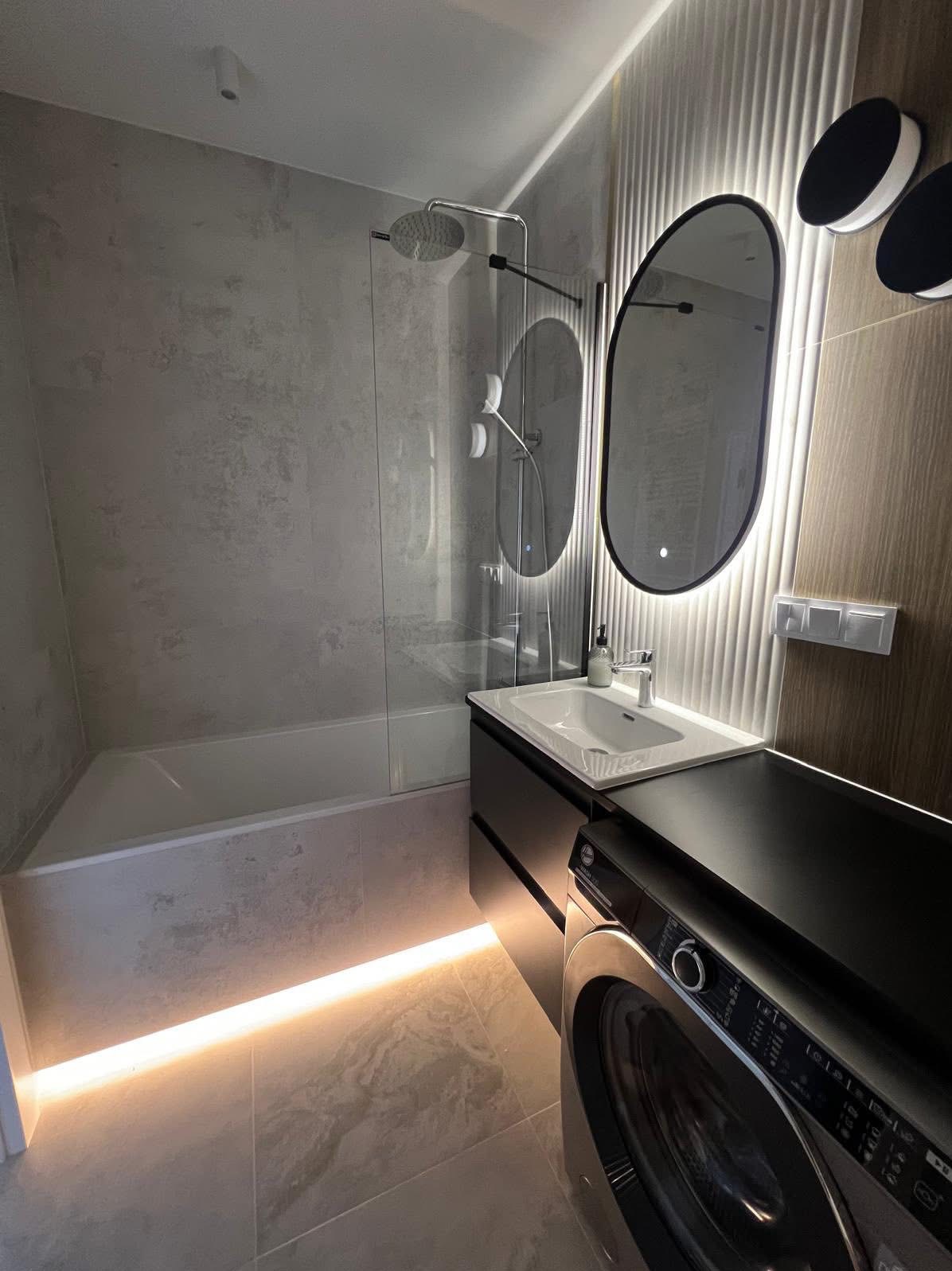 Nowoczesna łazienka z podświetlaną podłogą, owalnym lustrem z ledami i pralką w zabudowie. Minimalistyczny design, szare płytki i drewniane akcenty.