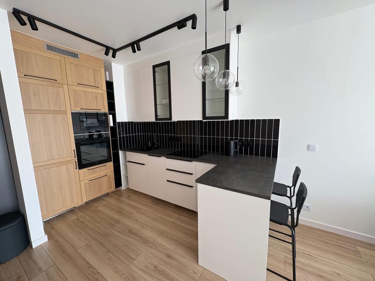 Nowoczesna kuchnia z drewnianymi i białymi szafkami, czarnym blatem i płytkami. Oświetlenie punktowe i wiszące lampy kuliste. Parkiet na podłodze.