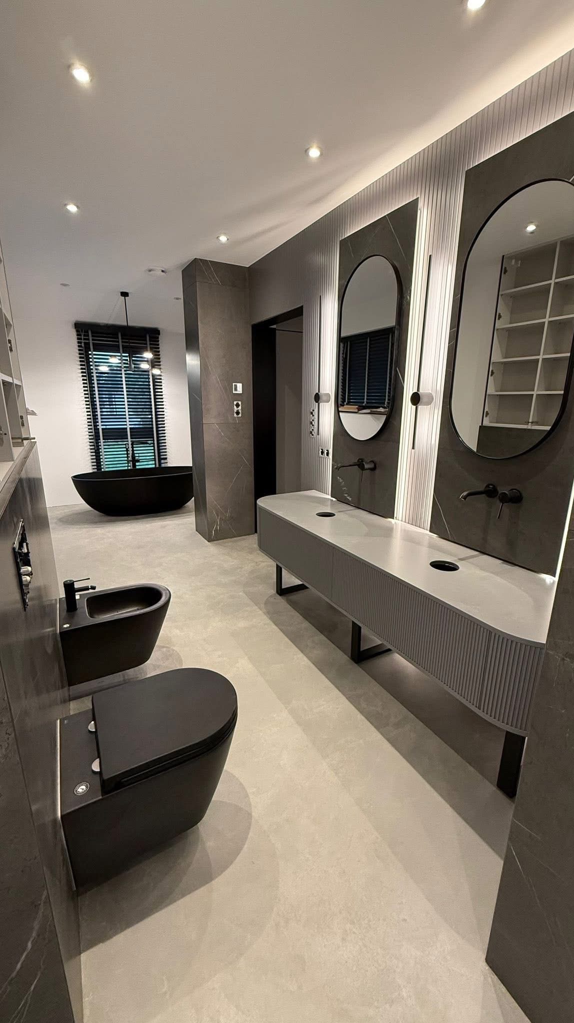 Nowoczesna łazienka w odcieniach szarości i czerni w Warszawie. Uwagę przykuwa wolnostojąca wanna i podwójna umywalka z lustrami w owalnych ramach.