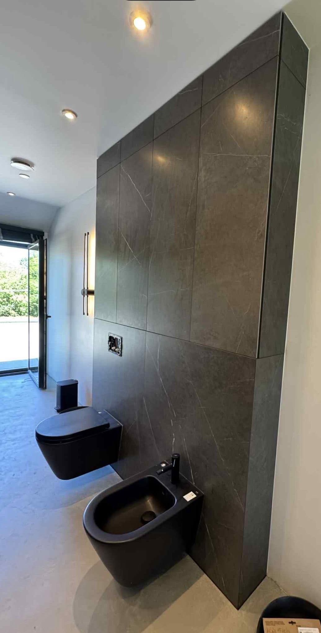 Nowoczesna łazienka z czarną armaturą i ścianą wyłożoną ciemnymi płytkami imitującymi marmur. Minimalistyczny design, oświetlenie punktowe i otwarte drzwi balkonowe.