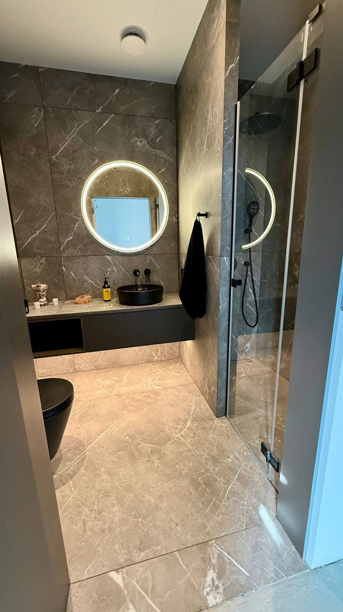 Nowoczesna łazienka z marmurowymi płytkami, okrągłym lustrem z podświetleniem LED i czarną armaturą. Kabina prysznicowa ze szklanymi drzwiami.