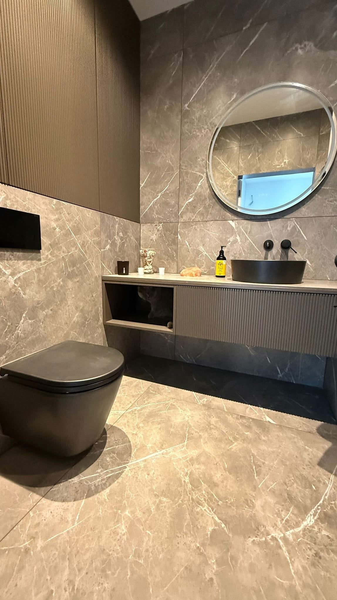 Elegancka łazienka w szarościach z marmurowymi płytkami, owalnym lustrem i podwieszaną toaletą. W szafce pod umywalką ukryty kot. Nowoczesny design.