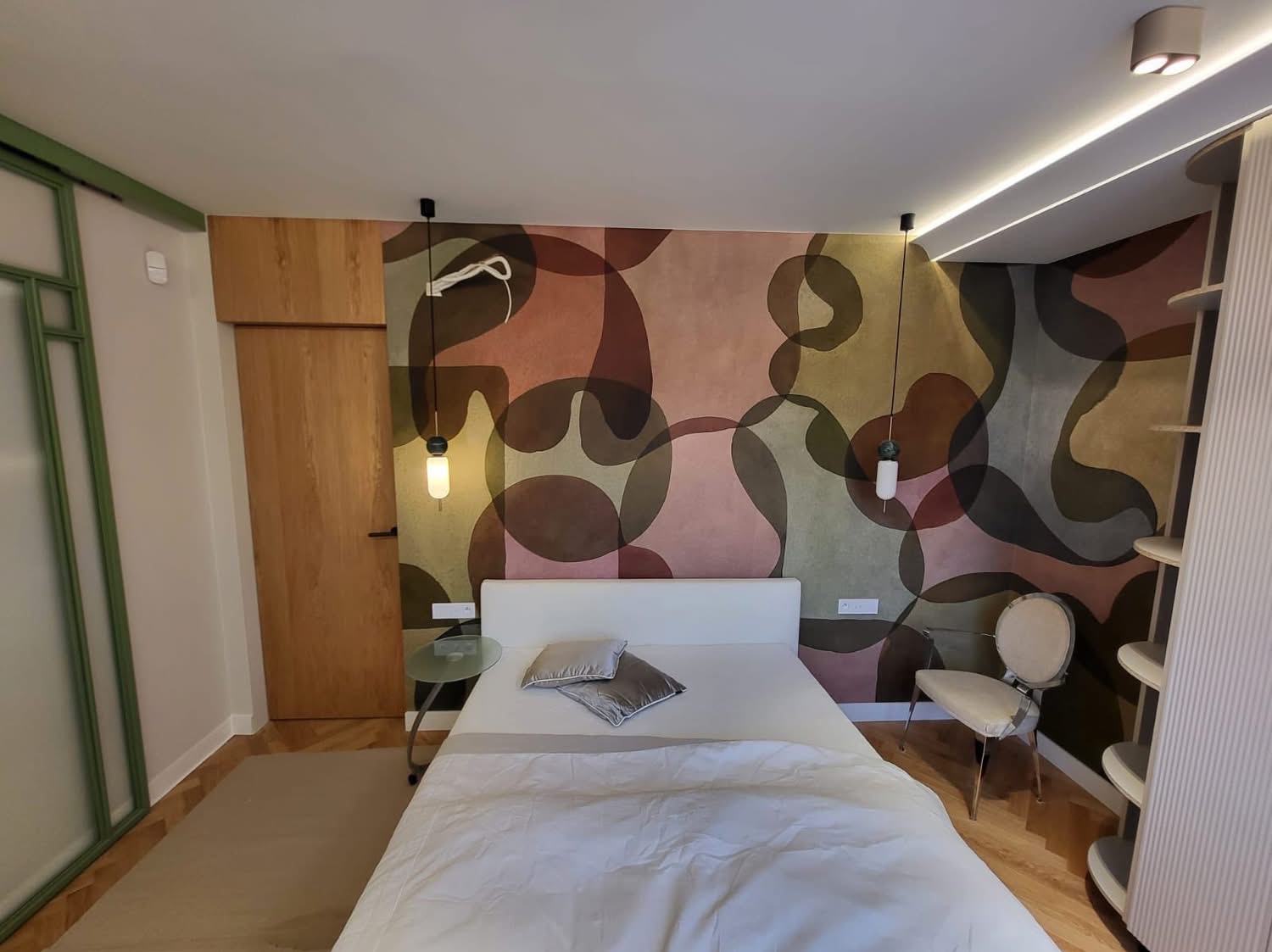 Sypialnia z łóżkiem na tle ściany z abstrakcyjnym wzorem. Wiszące lampy po bokach łóżka, drewniane drzwi i regał w tle. Jasne, nowoczesne wnętrze.