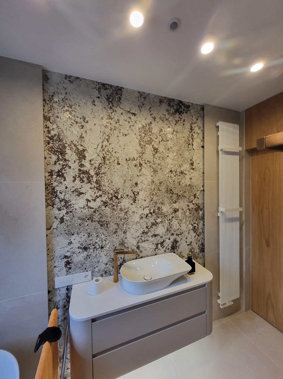 Nowoczesna łazienka z umywalką nablatową, złotą armaturą i dekoracyjną ścianą z płytek imitujących marmur. Grzejnik drabinkowy i drewniane drzwi w tle.