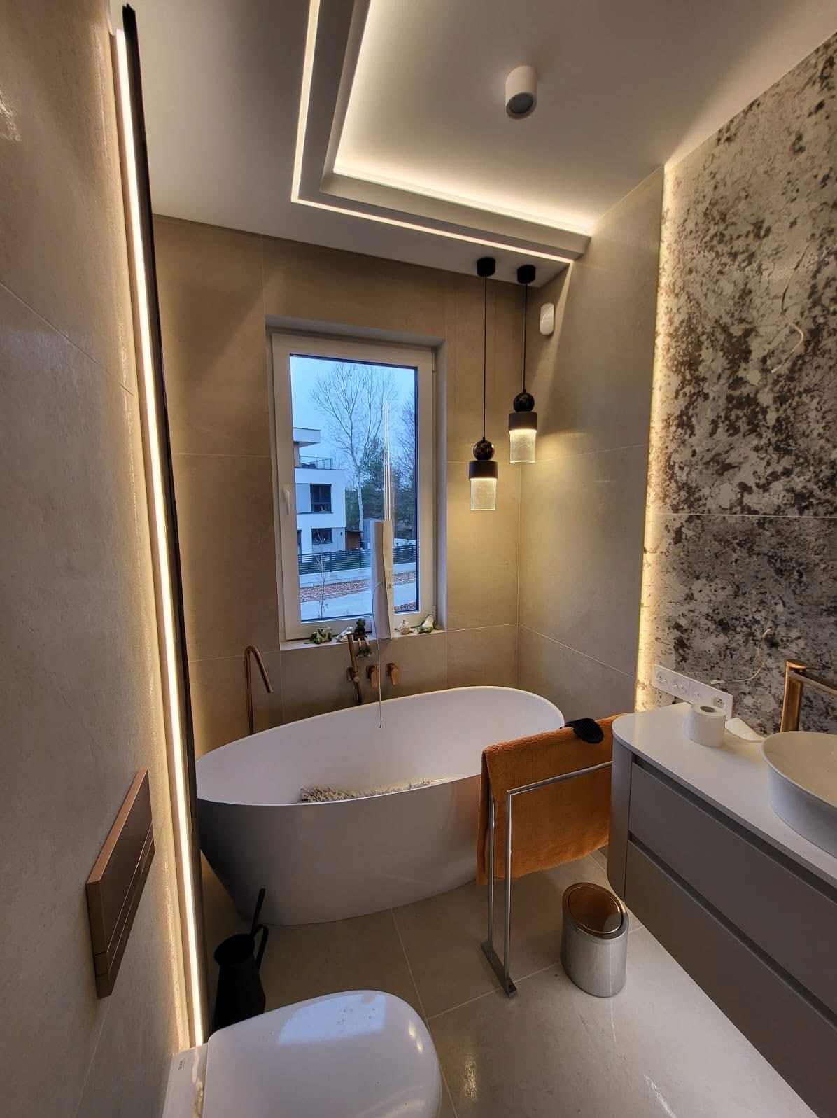 Nowoczesna łazienka z wolnostojącą wanną, beżowymi płytkami i dekoracyjną ścianą. Oświetlenie LED podkreśla minimalistyczny design i dodaje elegancji.