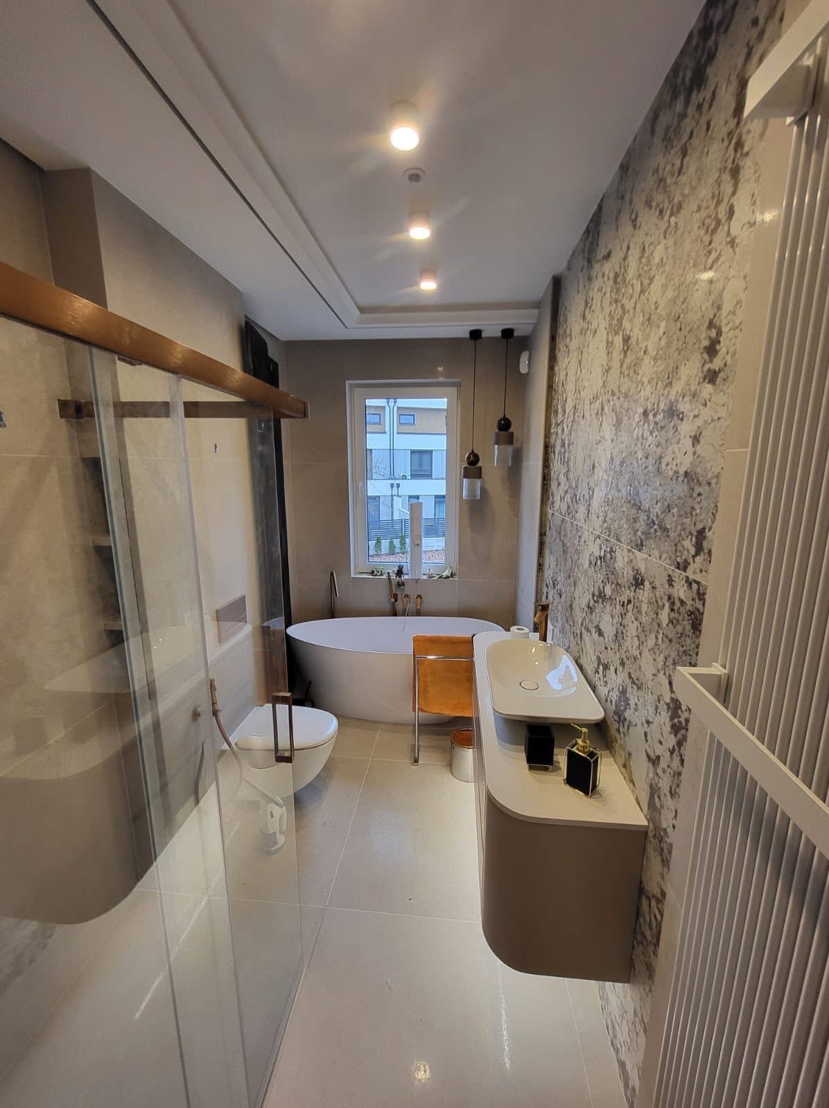 Nowoczesna łazienka z owalną wanną, kabiną prysznicową ze złotymi detalami i umywalką na szafce. Dekoracyjna ściana z efektem kamienia. Widok na okno.