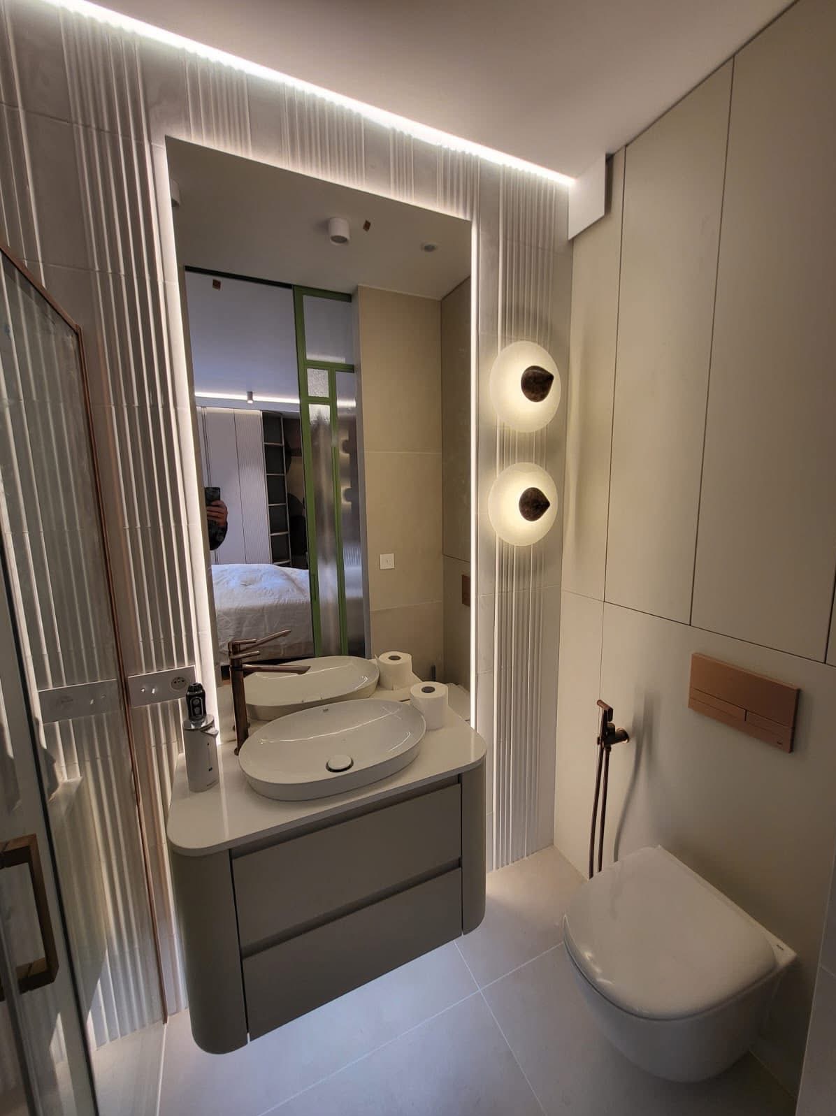 Nowoczesna łazienka z podświetlanym lustrem, umywalką na szafce i wiszącą toaletą. Minimalistyczny design z elementami dekoracyjnymi i subtelnym oświetleniem.