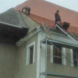 dach dek - Dwóch dekarzy na dachu budynku podczas wymiany pokrycia dachowego z czerwonej dachówki, widoczne rusztowanie i drabina.