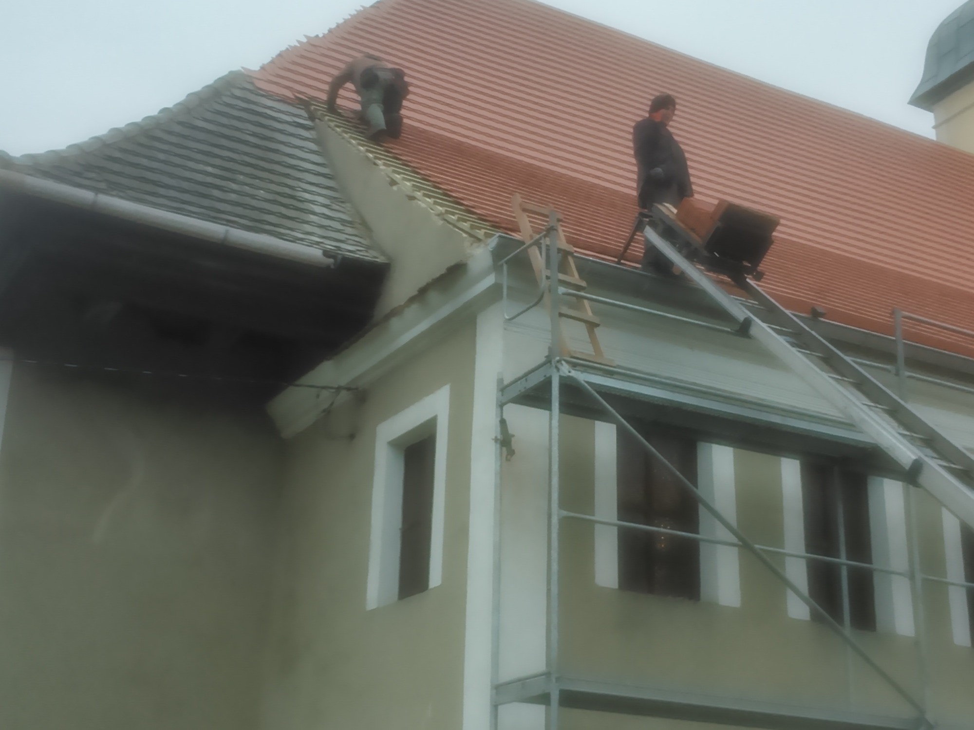 Dwóch dekarzy na dachu budynku podczas wymiany pokrycia dachowego z czerwonej dachówki, widoczne rusztowanie i drabina.