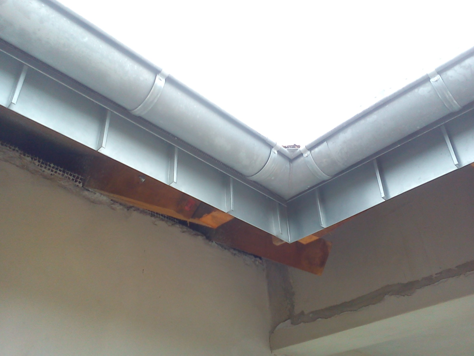 Szare, metalowe rynny narożne zamontowane na budynku, widoczne elementy konstrukcyjne dachu i ściany budynku.