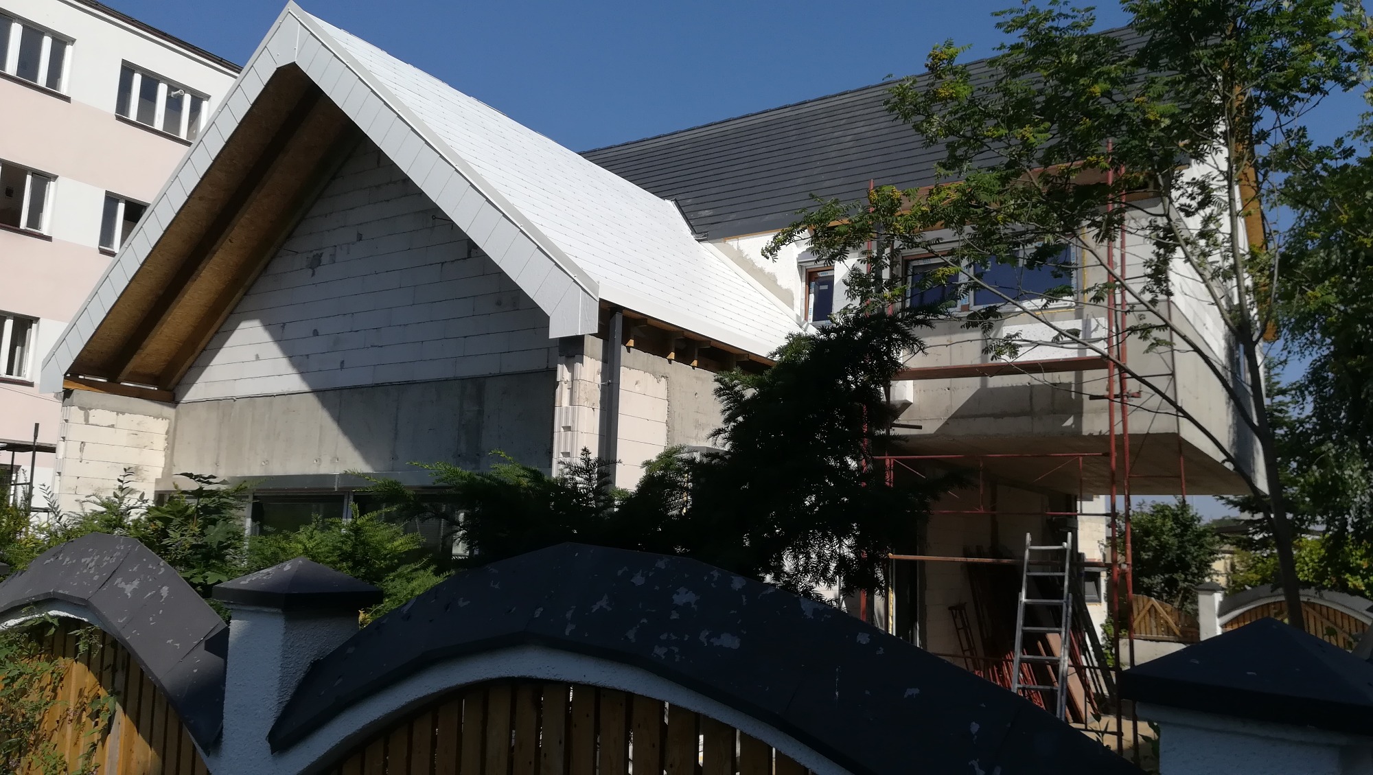 Dom w trakcie budowy z częściowo ukończonym białym dachem blaszanym i widocznym rusztowaniem, ogrodzony niskim płotem z czarnymi elementami.