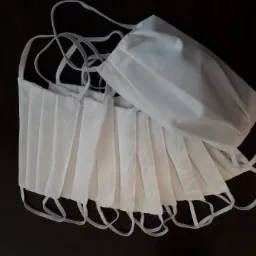 Stos białych, plisowanych maseczek ochronnych z elastycznymi gumkami na ciemnym, błyszczącym blacie.