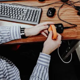 Mężczyzna w koszuli w paski podłącza pomarańczowy element do czarnego urządzenia elektronicznego na biurku z klawiaturą i myszą