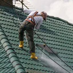 Pracownik w uprzęży, z żółtymi butami, czyści dach z zielonej dachówki za pomocą myjki ciśnieniowej, widoczny komin i antena.