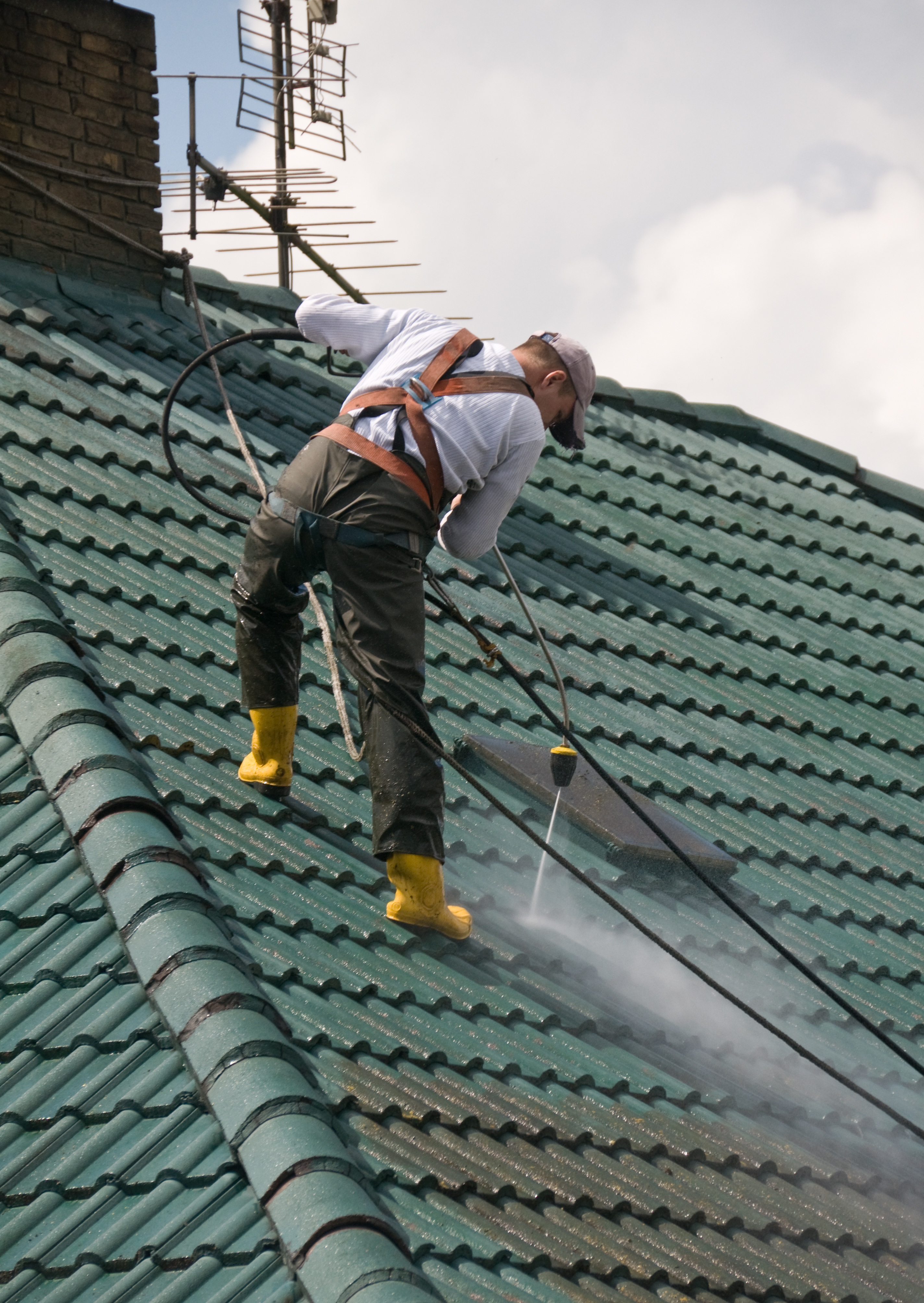 Pracownik w uprzęży, z żółtymi butami, czyści dach z zielonej dachówki za pomocą myjki ciśnieniowej, widoczny komin i antena.
