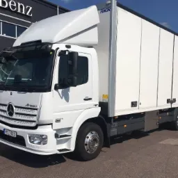 Biały samochód ciężarowy Mercedes-Benz Atego z zabudową kontenerową Fokor, zaparkowany przed budynkiem z logo Mercedes-Benz, widok z boku.