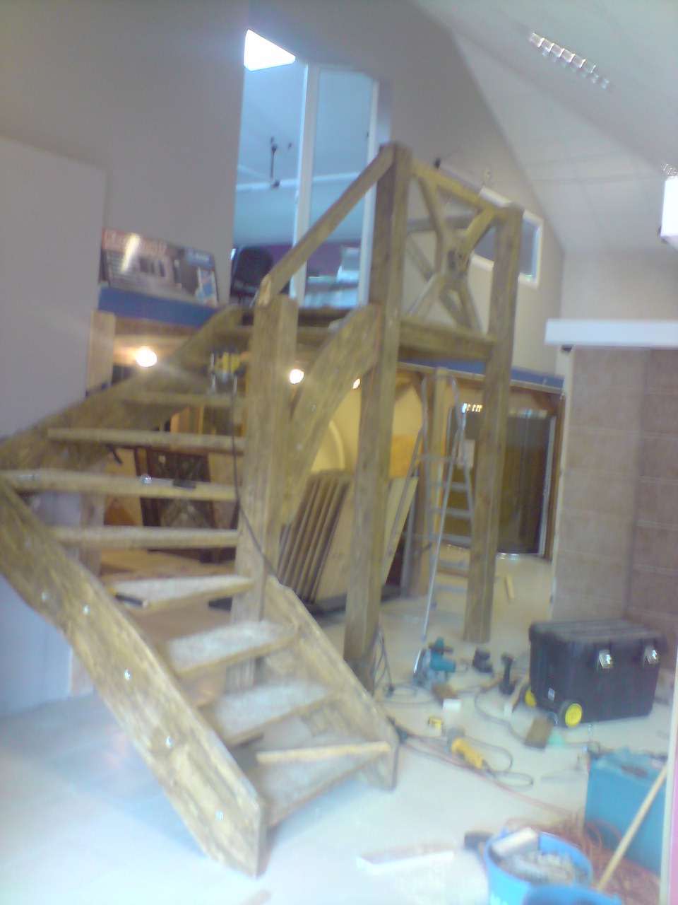 Drewniane schody w trakcie montażu, widoczne narzędzia i materiały budowlane, surowe wykończenie, w tle drabina i skrzynia narzędziowa.