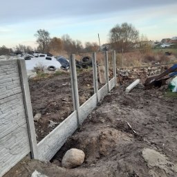 Usługi ziemno-inżynieryjne Kamil Bała - Montaż prefabrykowanego ogrodzenia betonowego w Rozdrażewie; widoczne słupki i panele z imitacją kamienia, ziemia po wykopach, w tle składowisko opon.