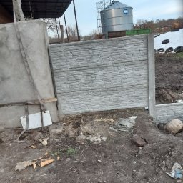 Usługi ziemno-inżynieryjne Kamil Bała - Fragment nowo postawionego, prefabrykowanego, betonowego ogrodzenia o fakturze kamienia, z widocznym słupkiem, na tle ziemi i zbiornika na zboże.