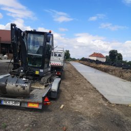 Usługi ziemno-inżynieryjne Kamil Bała - Mini koparka na lawecie obok świeżo wylanego fundamentu pod budynek, widoczne wykopane rowy i opony zabezpieczające.