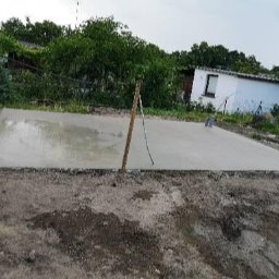 Usługi ziemno-inżynieryjne Kamil Bała - Świeżo wylany fundament z widocznymi śladami po narzędziach do rozprowadzania betonu, drewniany palik wbity w róg, w tle budynek gospodarczy i zieleń.