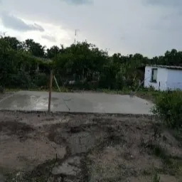 Świeżo wylana, mokra płyta betonowa na gruncie, widoczne narzędzia do rozprowadzania betonu, w tle budynek gospodarczy i drzewa pod pochmurnym niebem.