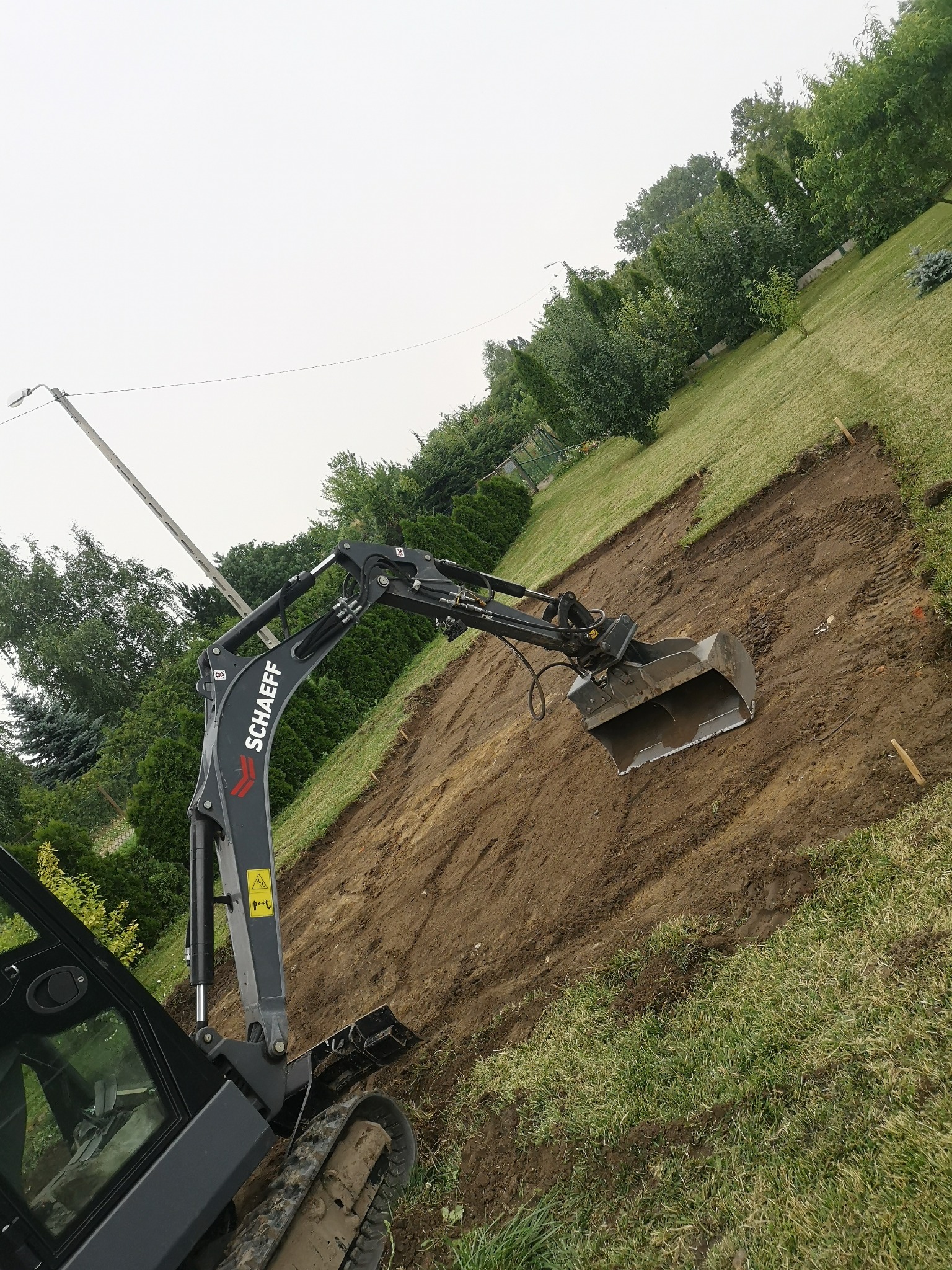 Czarny minikoparka Schaeff z łyżką, w trakcie prac ziemnych przy niwelacji terenu pod trawnik, widoczne ślady gąsienic i drewniane paliki wyznaczające obszar.