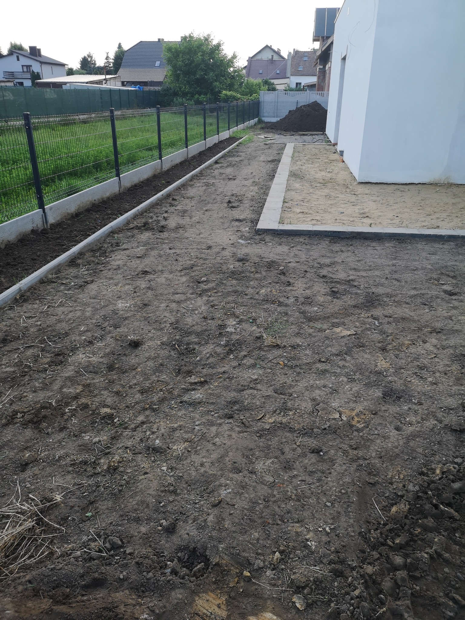 Przygotowany teren pod trawnik z obrzeżami betonowymi i zamontowanym ogrodzeniem panelowym, w tle widoczny dom w stanie surowym.