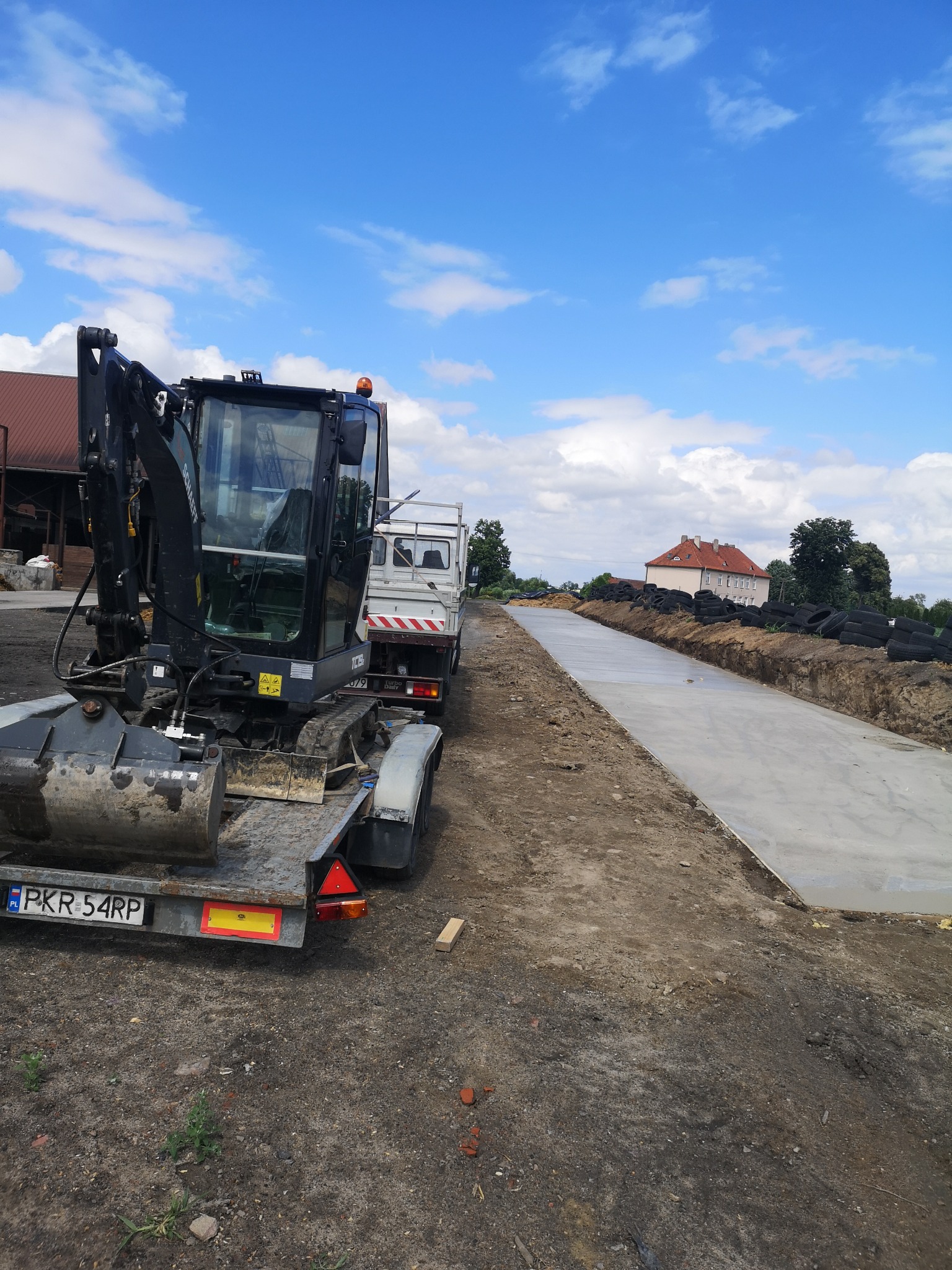 Mini koparka na lawecie obok świeżo wylanego fundamentu pod budynek, widoczne wykopane rowy i opony zabezpieczające.