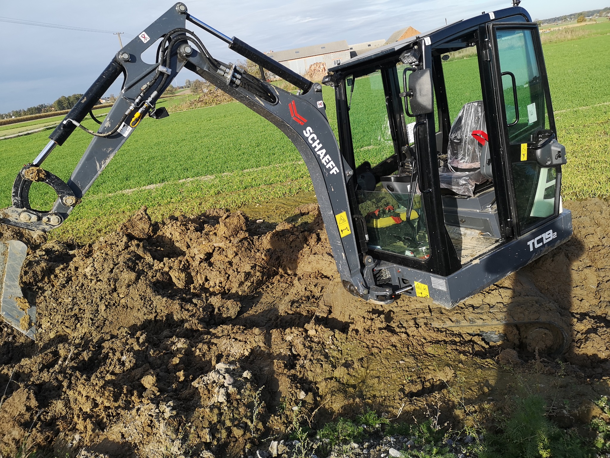 Czarny minikoparka Schaeff TC19 w trakcie pracy, z widoczną łyżką w ziemi i kabiną z plastikowymi osłonami na siedzeniu, na tle zielonego pola i budynku gospodarczego.