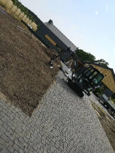 Mała koparka gąsienicowa na placu budowy, obok ułożona kostka brukowa, a dalej teren z brązową ziemią. W tle widoczny budynek z ciemną elewacją i jasną stolarką okienną.