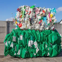 Dwie sprasowane bele odpadów: dolna z zielonych plastikowych kanistrów z etykietami ostrzegawczymi, górna z różnokolorowych, zgniecionych plastikowych opakowań, na tle betonowego muru i błękitnego...